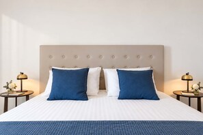 Uma cama bem arrumada com cabeceira estofada, duas mesas de cabeceira com abajures e um cobertor azul.