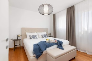 Une chambre à coucher avec un lit, des tables de chevet, un luminaire suspendu, une fenêtre avec des rideaux et un plancher en bois.