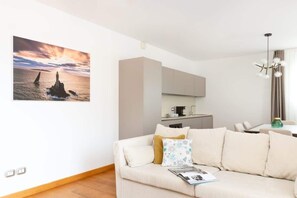 Un salon avec un canapé, un magazine, un tableau représentant un phare, et un coin cuisine à l’arrière-plan.