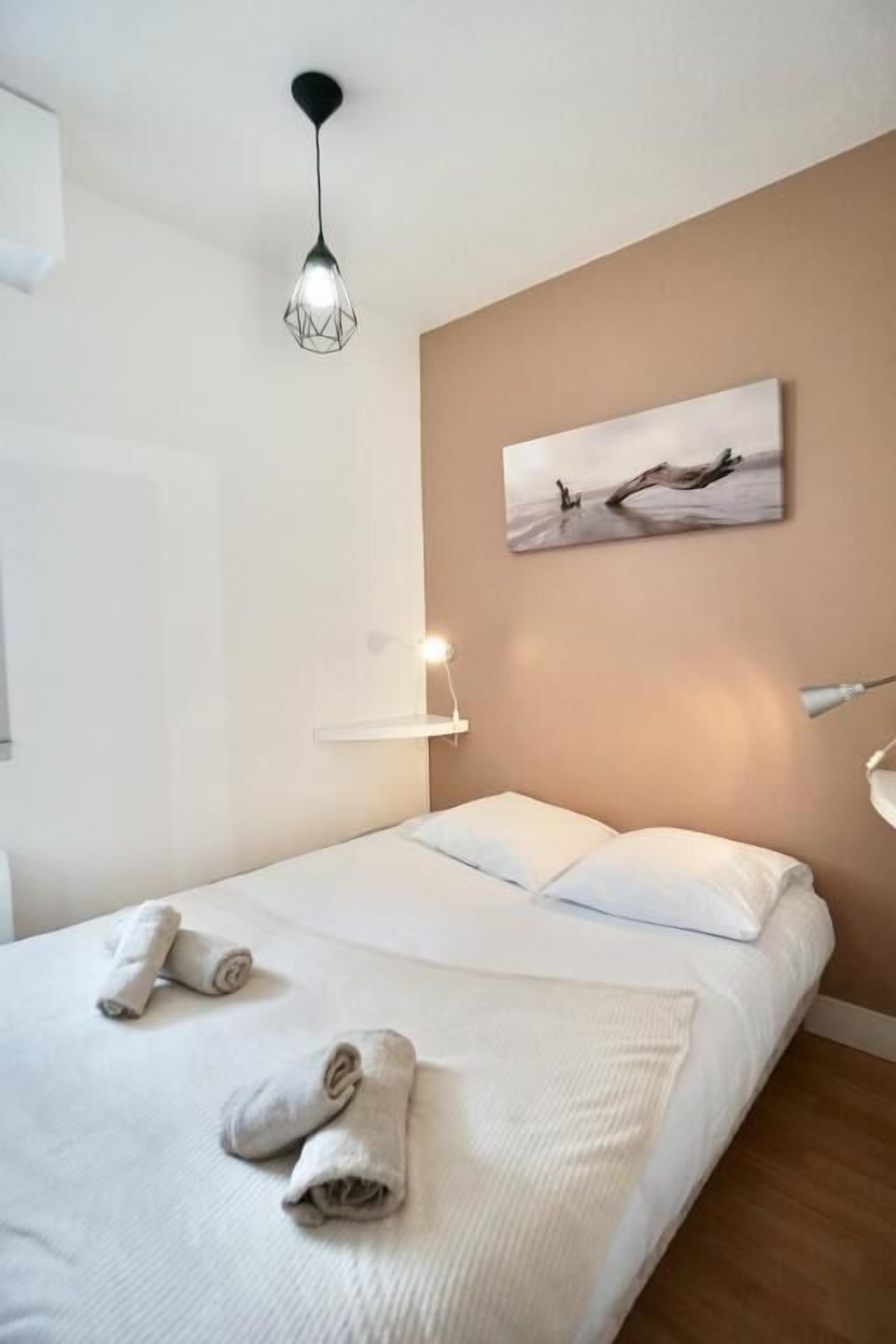 A hotel room with a bed, towels, a painting on the wall, a hanging light fixture, and a bedside table.