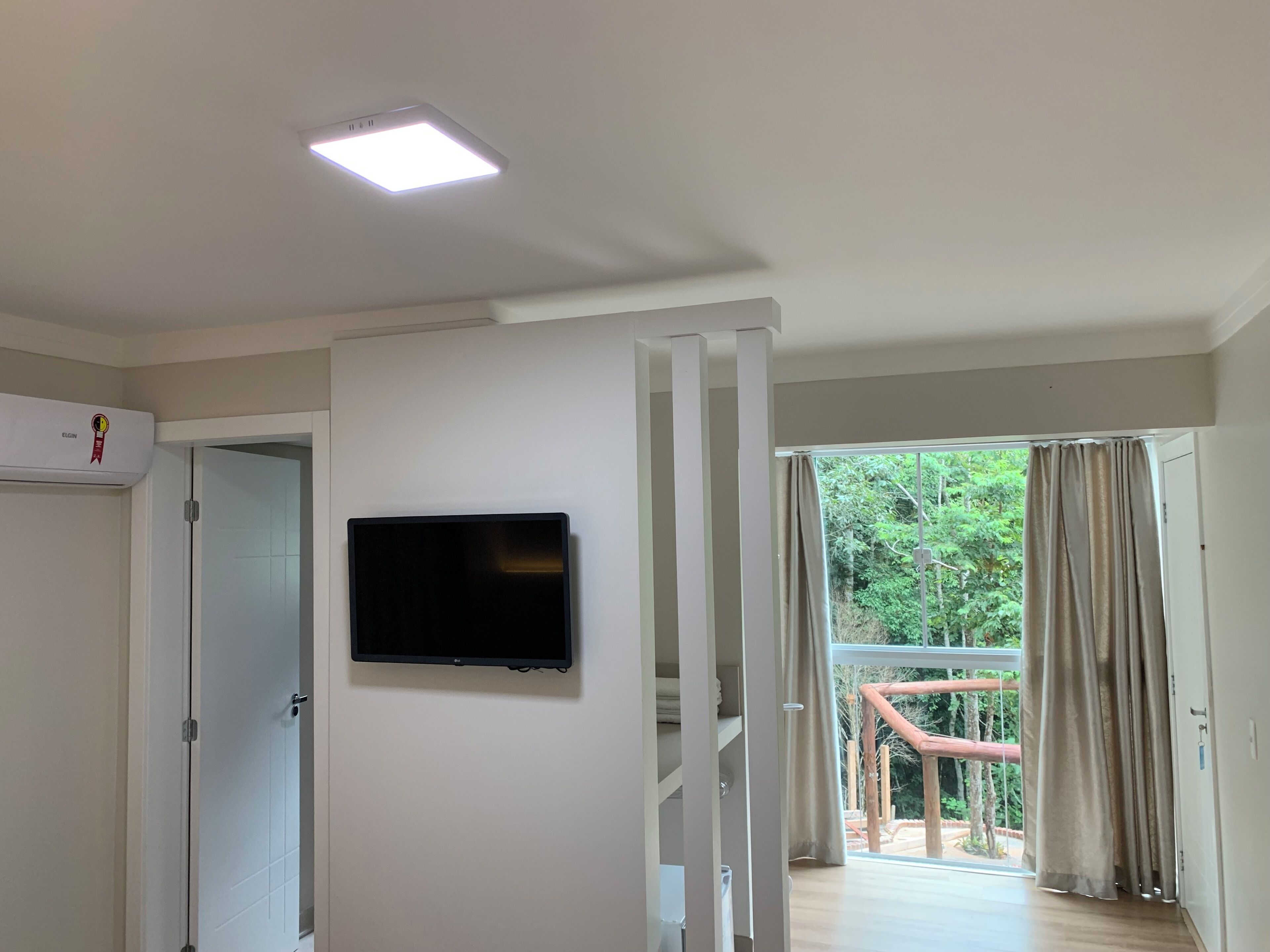 A room with a flat-screen TV mounted on a white wall, a ceiling light, and a door leading to an outdoor area with a wooden deck and greenery.