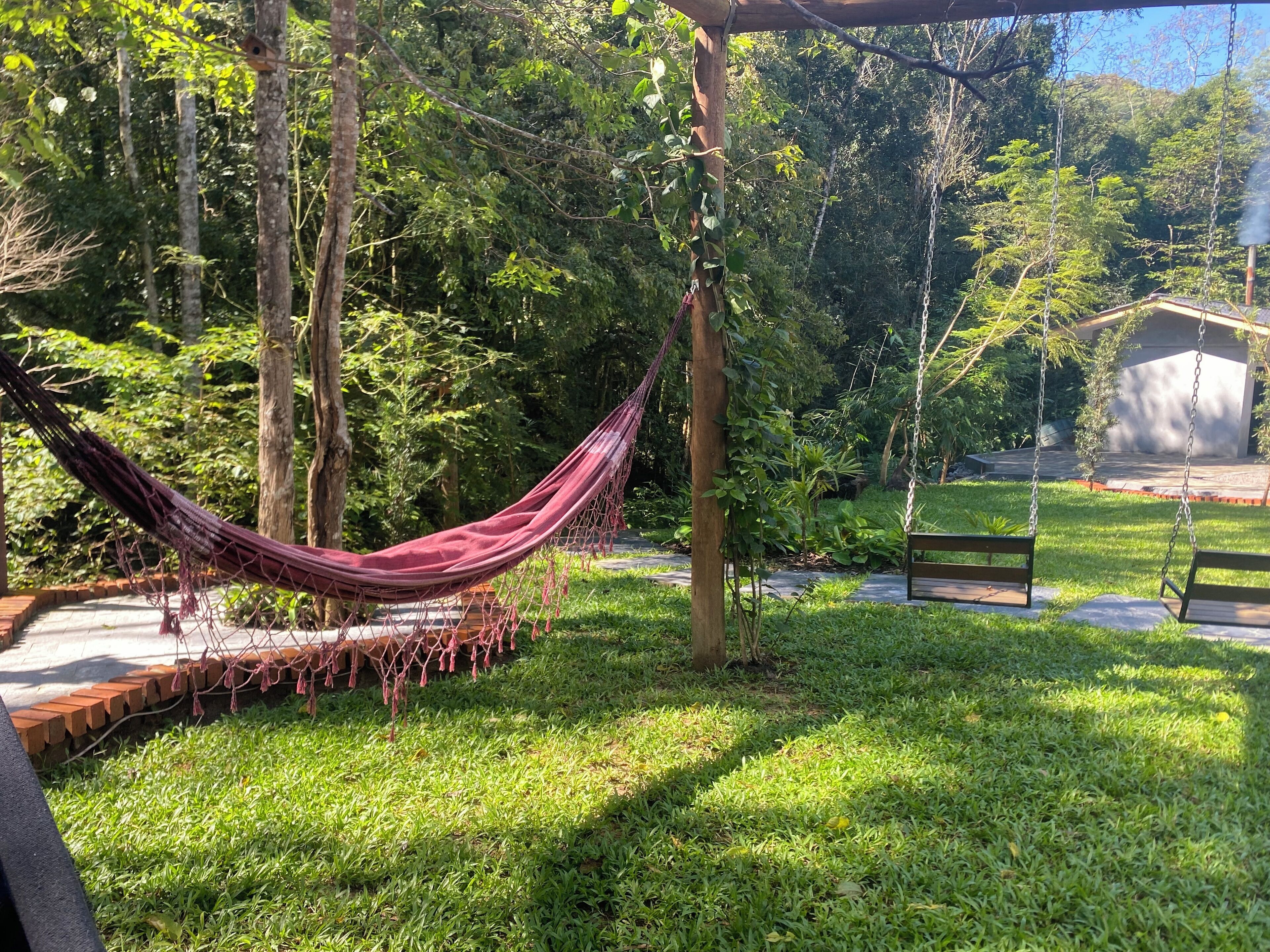 Una hamaca atada entre árboles en un jardín exuberante, con un banco y un cobertizo pequeño.