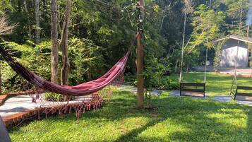Una hamaca atada entre árboles en un jardín exuberante, con un banco y un cobertizo pequeño.