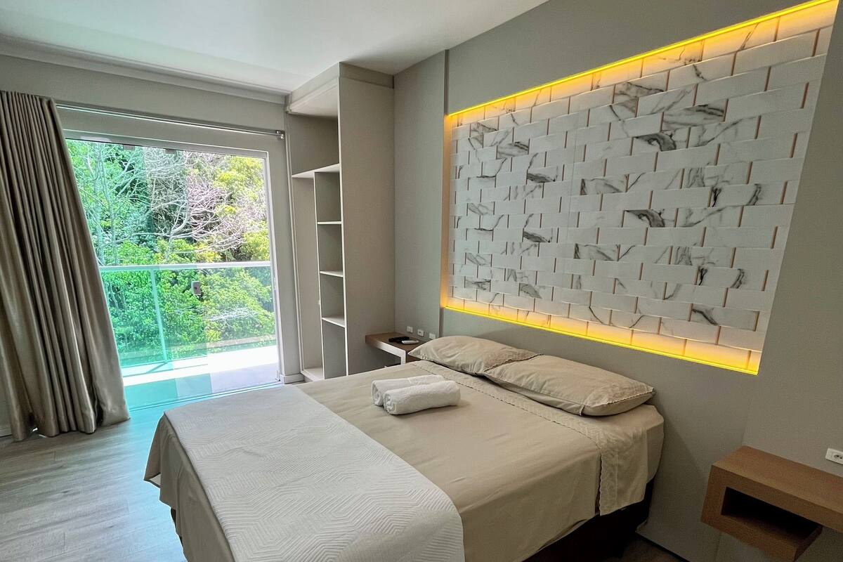 Ein modernes Schlafzimmer mit einem großen Bett, einer Wand im Marmoroptik, einem kleinen Nachttisch und Blick ins Grüne durch eine Schiebetür.