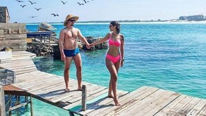 Um píer de madeira se estende sobre um mar azul e límpido, com um casal de mãos dadas.
