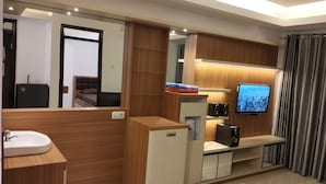 Un baño moderno con tocador, lavamanos y espejo. Hay un televisor montado en la pared y un mueble pequeño debajo.