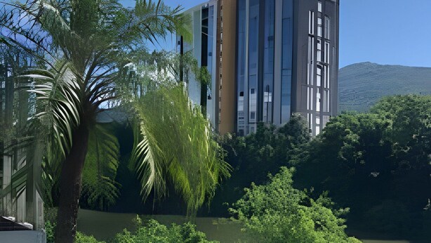 A modern building with a glass facade, surrounded by lush greenery and a body of water. - Saint Sebastian ap113-stúdio para casal (Jaragua do Sul)