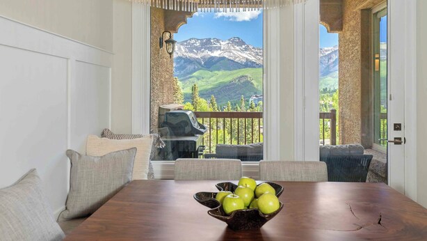 Ein Essbereich mit einem Holztisch, einer Schüssel grüner Äpfel und Blick auf Berge durch eine offene Tür.