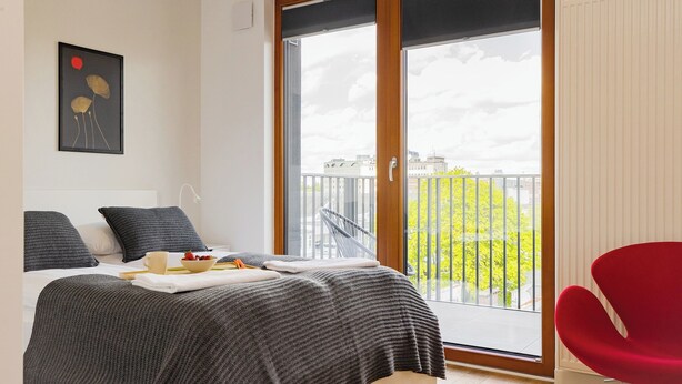A modern bedroom with a large sliding glass door leading to a balcony, a red chair, and a framed artwork on the wall. - ShortStayPoland Zdziechowskiego B151 (Warsaw)