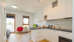 A modern kitchen with white cabinets, a black countertop, and stainless steel appliances. There is a red chair and a small table with a plant. - ShortStayPoland Zdziechowskiego B151 (Warsaw)