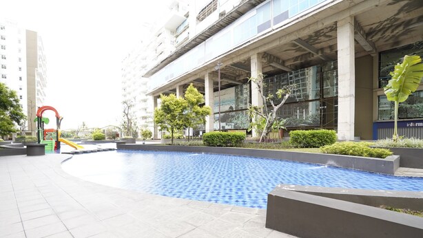 Una piscina con borde de baldosas, rodeada de vegetación y un edificio moderno con amplias ventanas y columnas.