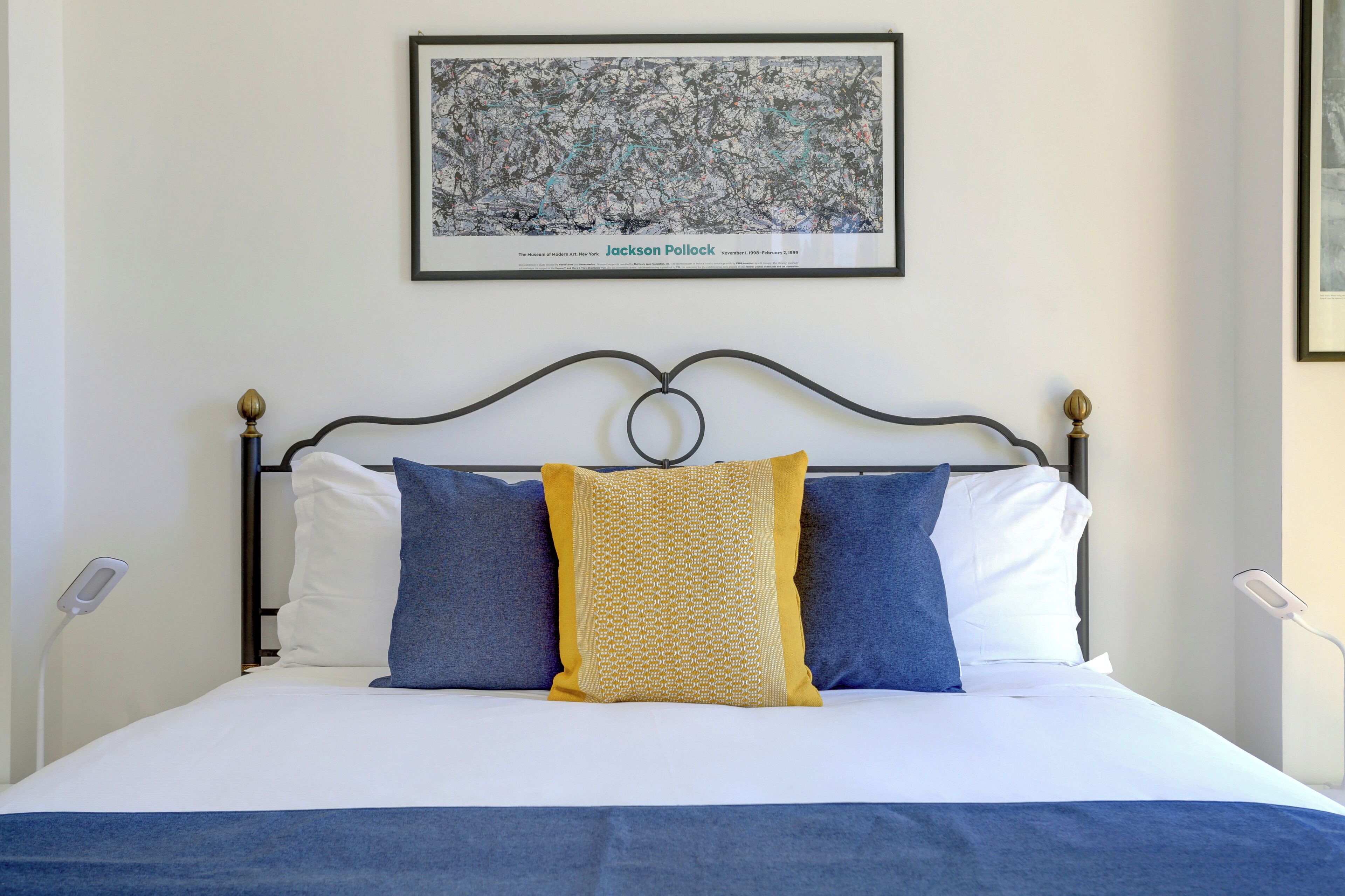 Una cama bien hecha con edredón blanco y azul, una almohada estampada de color amarillo y otra almohada azul. Un cuadro enmarcado cuelga sobre la cama.