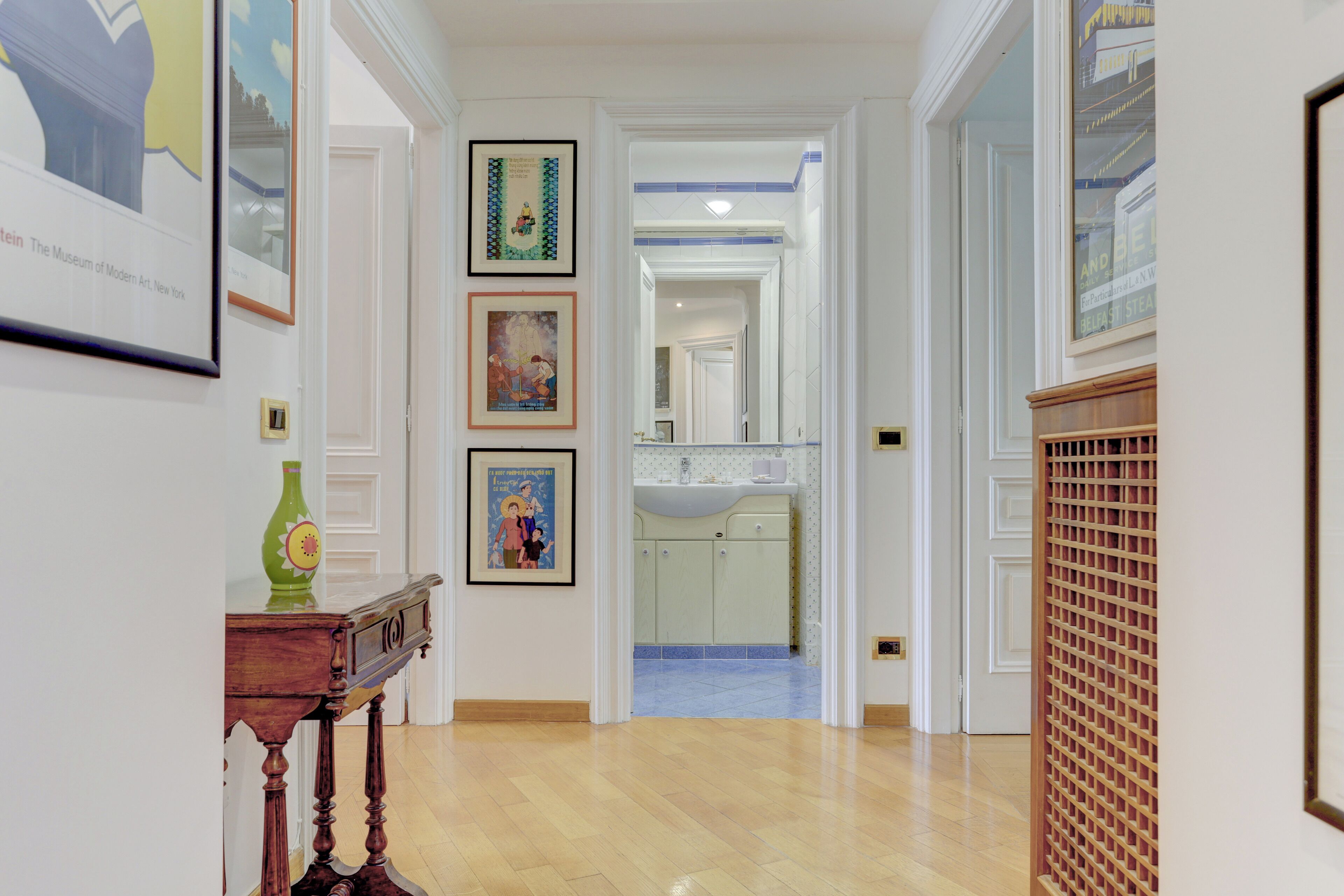 A hallway with framed artwork on the walls, a wooden table with a vase, and a view into a bathroom with a sink and mirror.