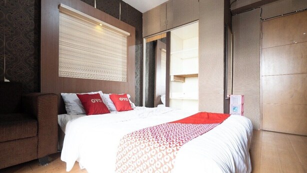 A hotel room with a bed, a sofa, a large window with blinds, and a wardrobe. - OYO 93945 The Suite Metro Apartemen By Sa (Bandung)