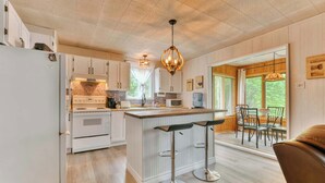 A modern kitchen with white cabinetry, a central island with bar stools, a built-in microwave, and a hanging light fixture. - Bord de lac | Chalet du Bonheur (Amherst)
