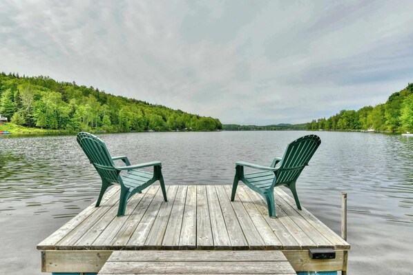 Eine Holzsteganlage mit zwei grünen Adirondack-Stühlen, die auf einen ruhigen See mit bewaldeten Hügeln blicken.