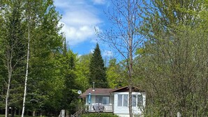 A single-story house with a visible deck, surrounded by a wooded area and a clear sky. - Bord de lac | Chalet du Bonheur (Amherst)