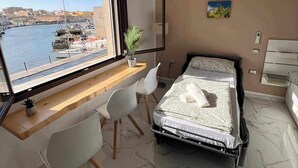 Ein modernes Schlafzimmer mit großem Fenster mit Blick auf eine Marina, einem Schreibtisch mit Stuhl, einem Bett und einer kleinen Pflanze.