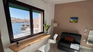 Ein modernes Wohnzimmer mit großem Fenster mit Blick auf eine Marina, einer Couch, zwei weißen Sesseln und einem kleinen Tisch mit einer Pflanze.