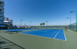 Una cancha de tenis con valla verde, faroles y cielo despejado.