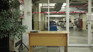 A reception desk with a potted plant, glass doors leading to a parking area with cars, and directional signs. - Capital O 93961 Apartement Sentraland By Helen (Karawang)