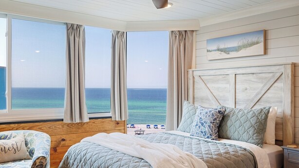 Un dormitorio con un ventanal grande con vistas al océano, cabecera de madera y una cama con muchas almohadas.