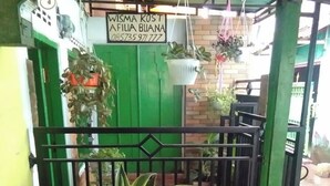 A green gate with a sign reading "WISMA KOST AFILIA BUANA" and a contact number. There are potted plants on a table and hanging from the gate. - Hotel O Wisma Kost & Penginapan Afilia Buana Syariah (Tulungagung)