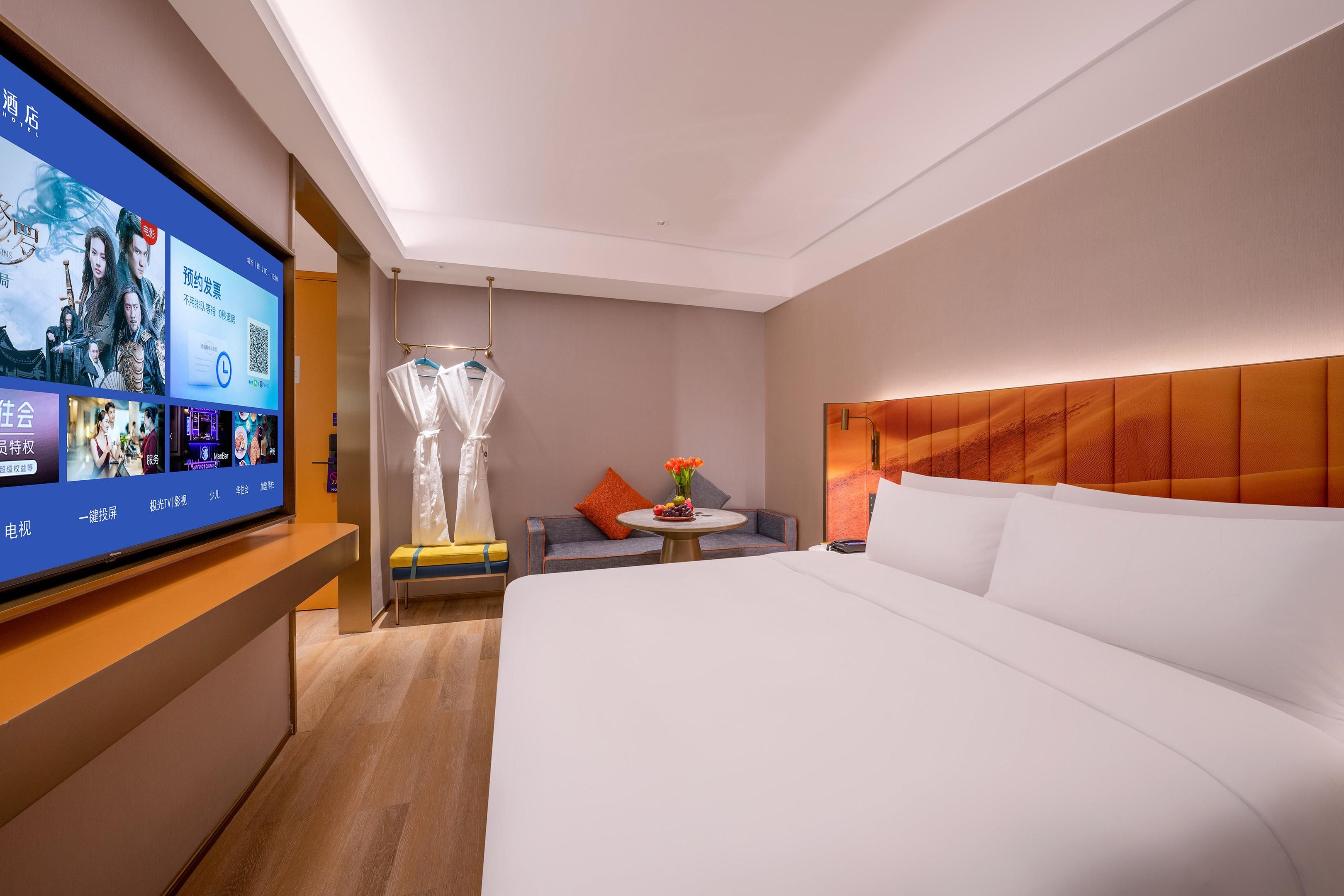 A hotel room with a large bed, a television, a small table with a vase, and a seating area with a chair and a sofa.