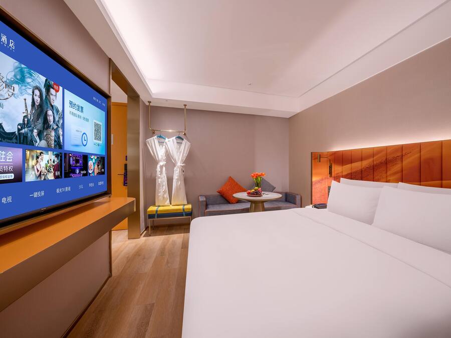 Habitación de hotel con una cama grande, televisión, una mesita con un florero y un rincón de descanso con una silla y un sofá.