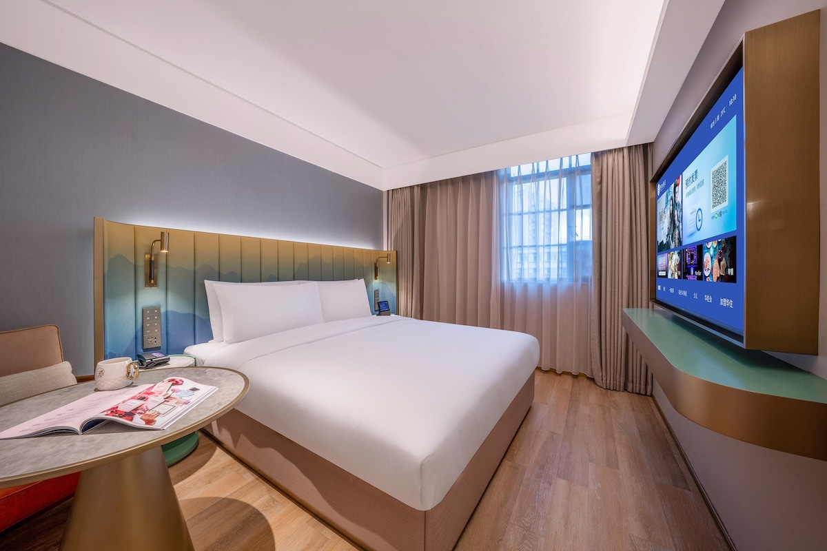 Une chambre d’hôtel avec un grand lit, une table de chevet avec un magazine, un téléviseur posé sur un meuble et une fenêtre avec des rideaux.