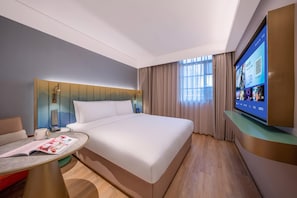Ein Hotelzimmer mit einem großen Bett, einem an der Wand befestigten Fernseher, einem Schreibtisch mit einer Zeitschrift und einem Fenster mit Vorhängen.