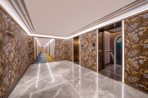 A hotel corridor with a marble floor, floral wallpaper, and a central elevator. - Manxin Hotel Yantai Binhai Plaza (Yantai)