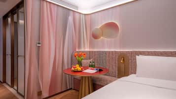 A hotel room with a bed, a small round table with a fruit bowl, a vase with flowers, and a patterned headboard.