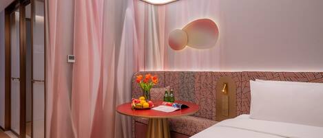 A hotel room with a bed, a small round table with a fruit bowl, a vase with flowers, and a patterned headboard.