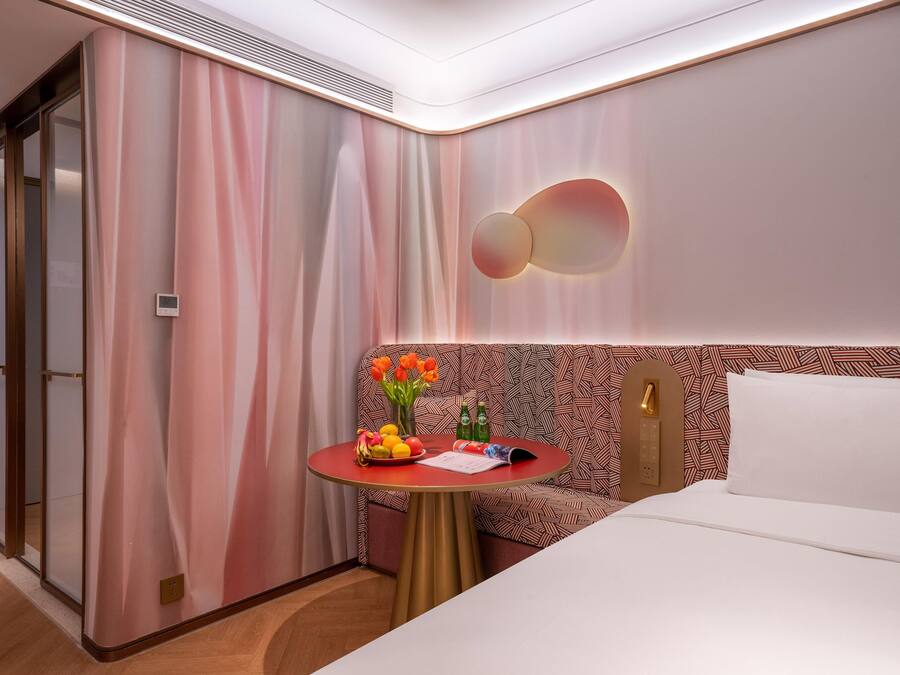 A hotel room with a bed, a small round table with a fruit bowl, a vase with flowers, and a patterned headboard.