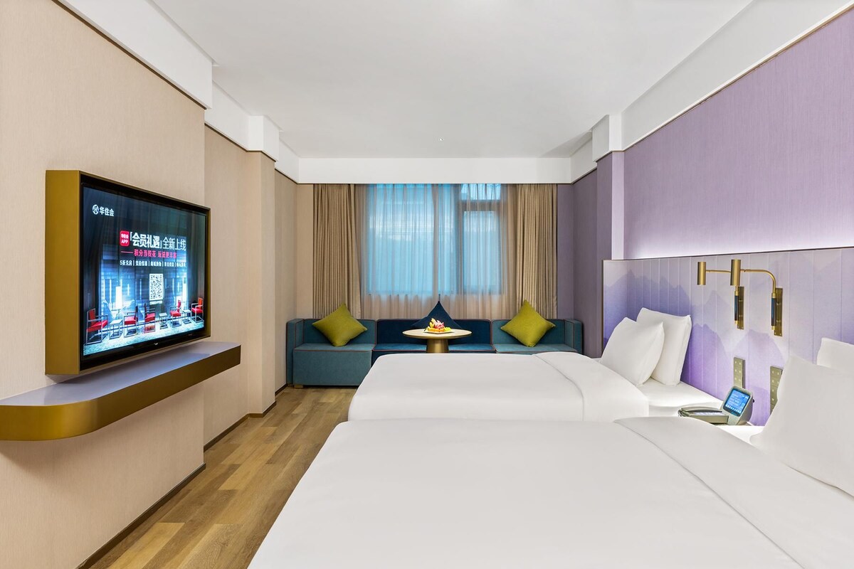 Une chambre d’hôtel avec un grand lit, un téléviseur fixé au mur, un coin salon avec des coussins et une fenêtre avec des rideaux.