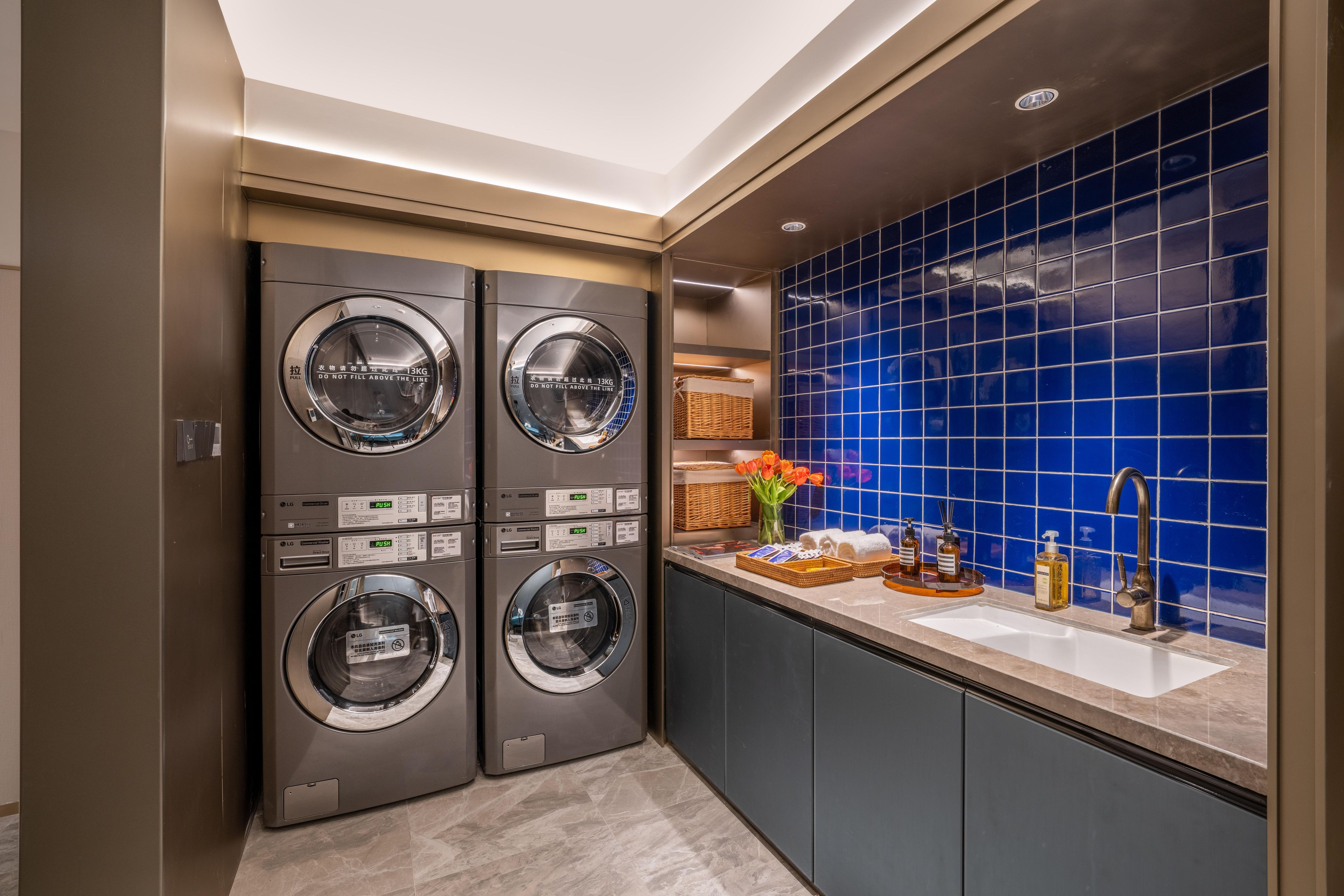 Un moderno lavadero con cuatro lavadoras comerciales LG, un lavabo con artículos de aseo y un salpicadero de azulejos azules.