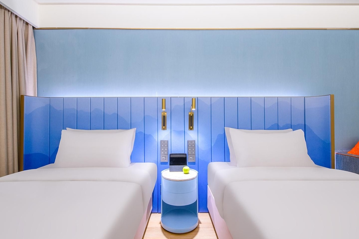 A hotel room with two beds, a bedside table with a lamp, and a wall-mounted control panel.