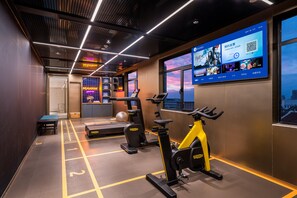 A modern gym with exercise equipment, a digital screen displaying information, and a city view through large windows.