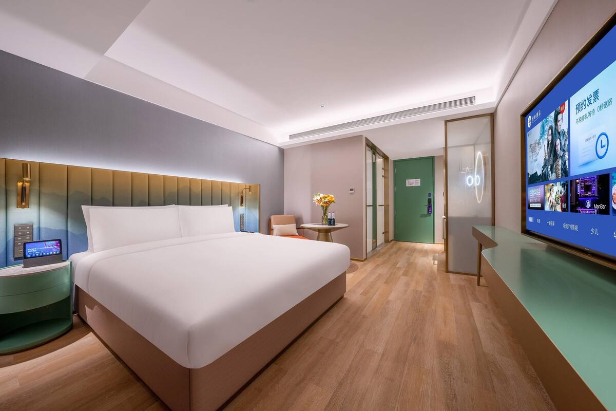 Ein modernes Hotelzimmer mit einem großen Bett, einem Nachttisch mit einem digitalen Gerät, einem kleinen runden Tisch mit einer Vase und einem großen Bildschirm, der Inhalte anzeigt.
