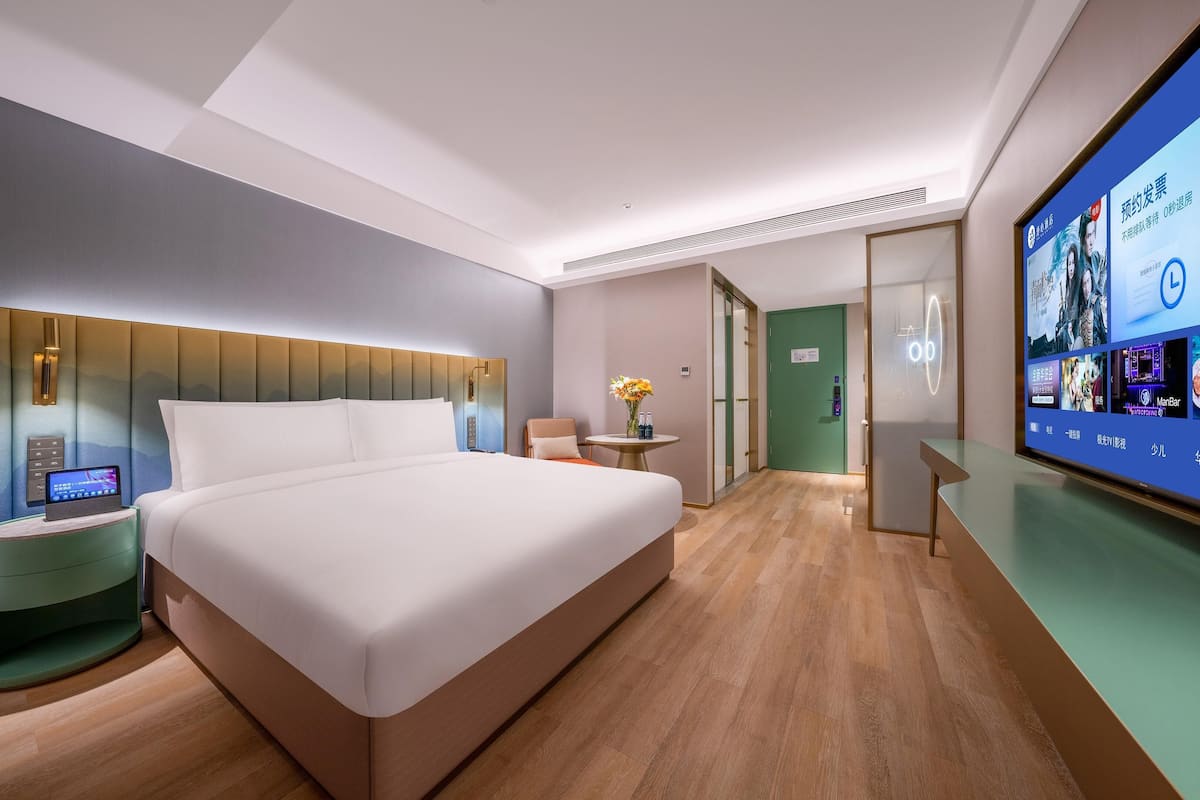 A modern hotel room with a large bed, a bedside table with a digital device, a small round table with a vase, and a large screen displaying content.