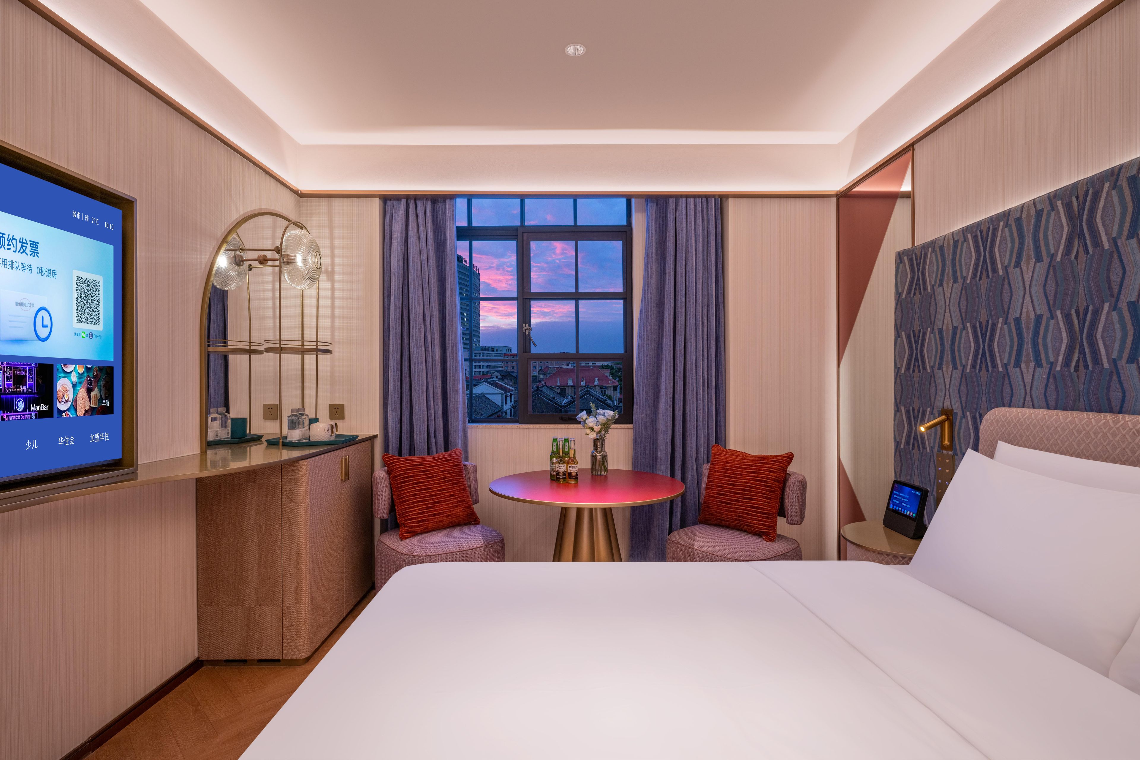 A hotel room with a bed, a desk with a computer, a window with a city view, and a small table with two chairs.