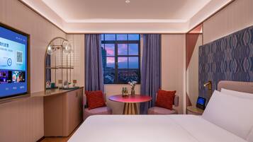 A hotel room with a bed, a desk with a computer, a window with a city view, and a small table with two chairs.