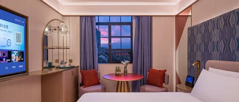 A hotel room with a bed, a desk with a computer, a window with a city view, and a small table with two chairs.