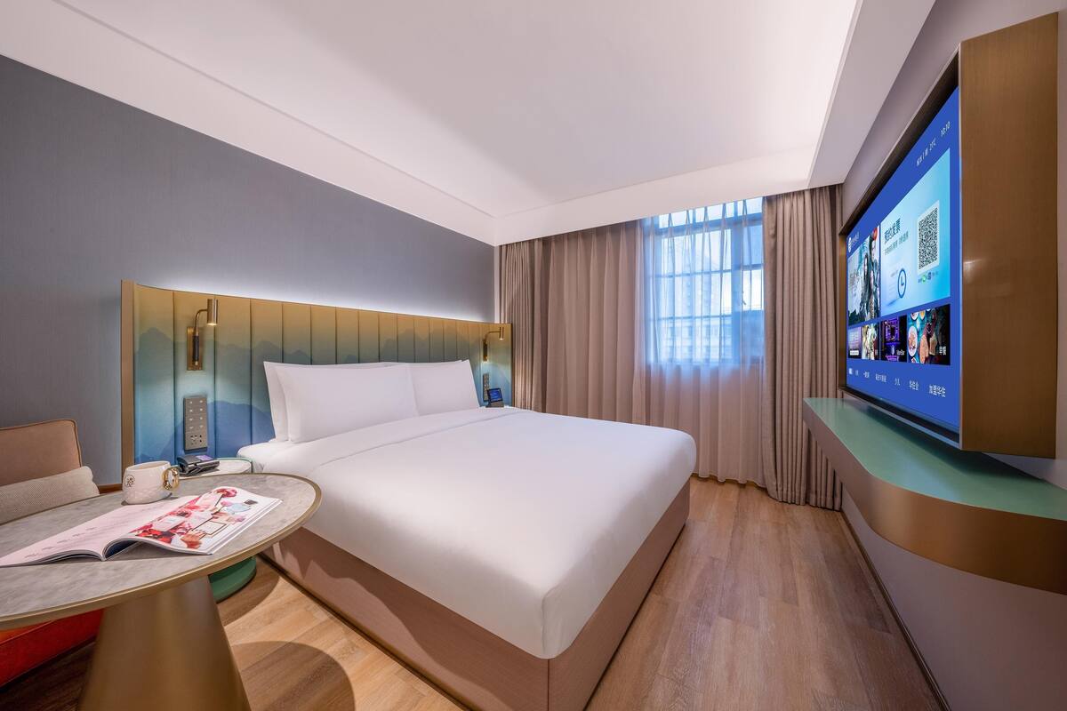 Ein Hotelzimmer mit einem großen Bett, einem Nachttisch mit einer Zeitschrift, einem Fernseher auf einem Standfuß und einem Fenster mit Vorhängen.