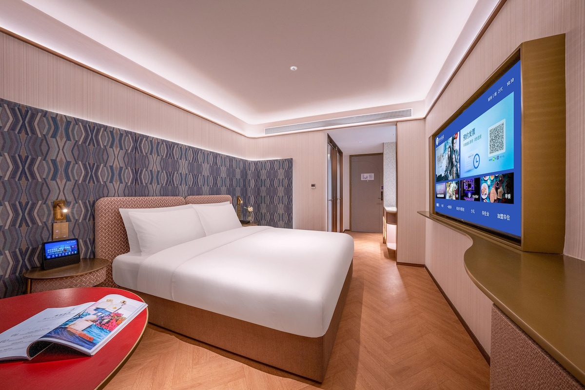 A hotel room with a bed, a TV on the wall, a bedside table with a lamp, and a magazine on the floor.