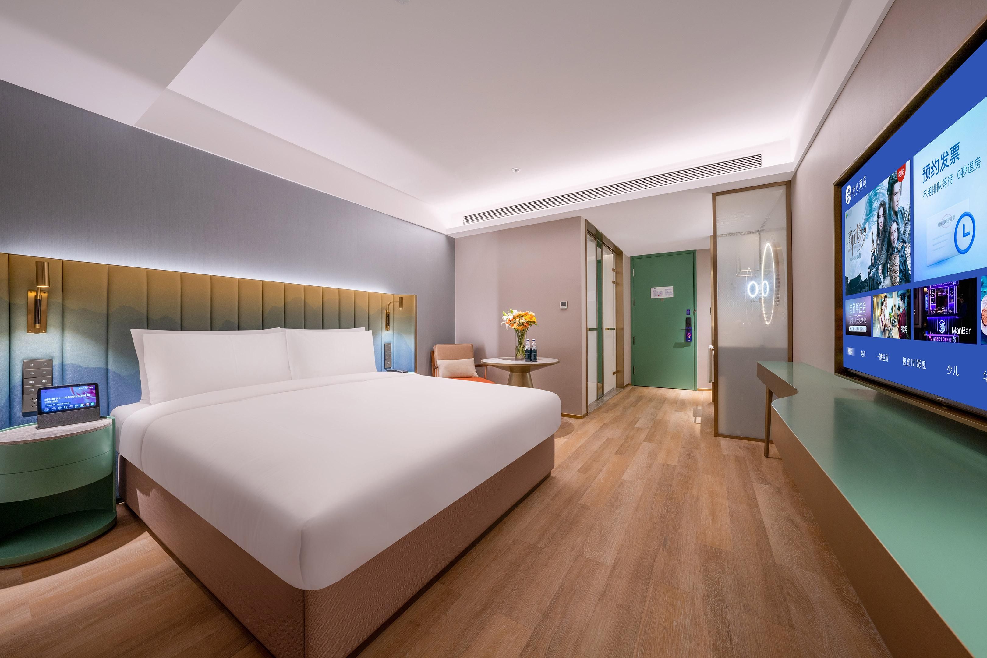 Una habitación de hotel moderna con una cama grande, una mesita de noche con un dispositivo digital, una pequeña mesa redonda con un florero y una pantalla grande mostrando contenido.