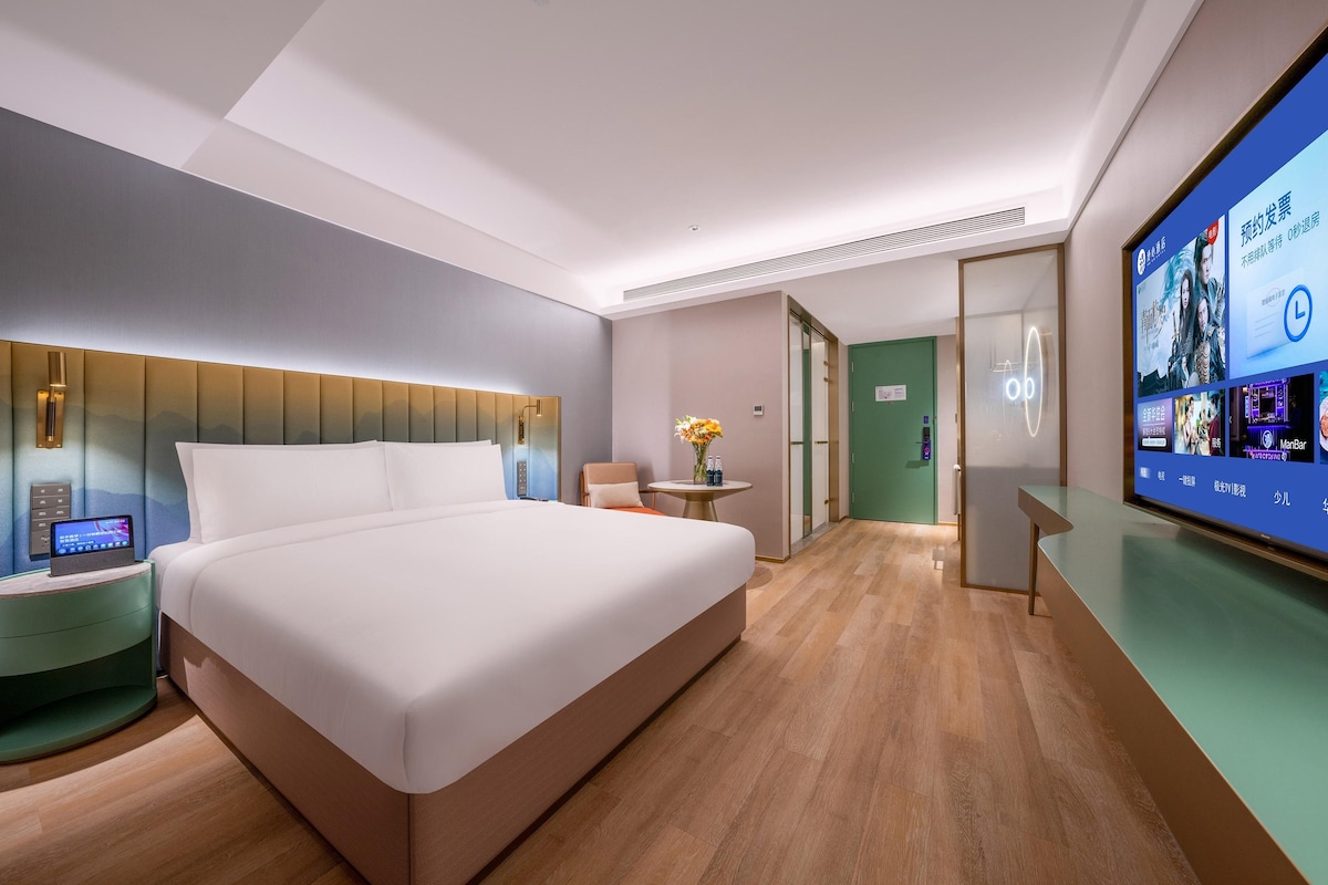 Une chambre d’hôtel moderne dotée d’un grand lit, d’une table de chevet avec un appareil numérique, d’une petite table ronde avec un vase et d’un grand écran diffusant du contenu.