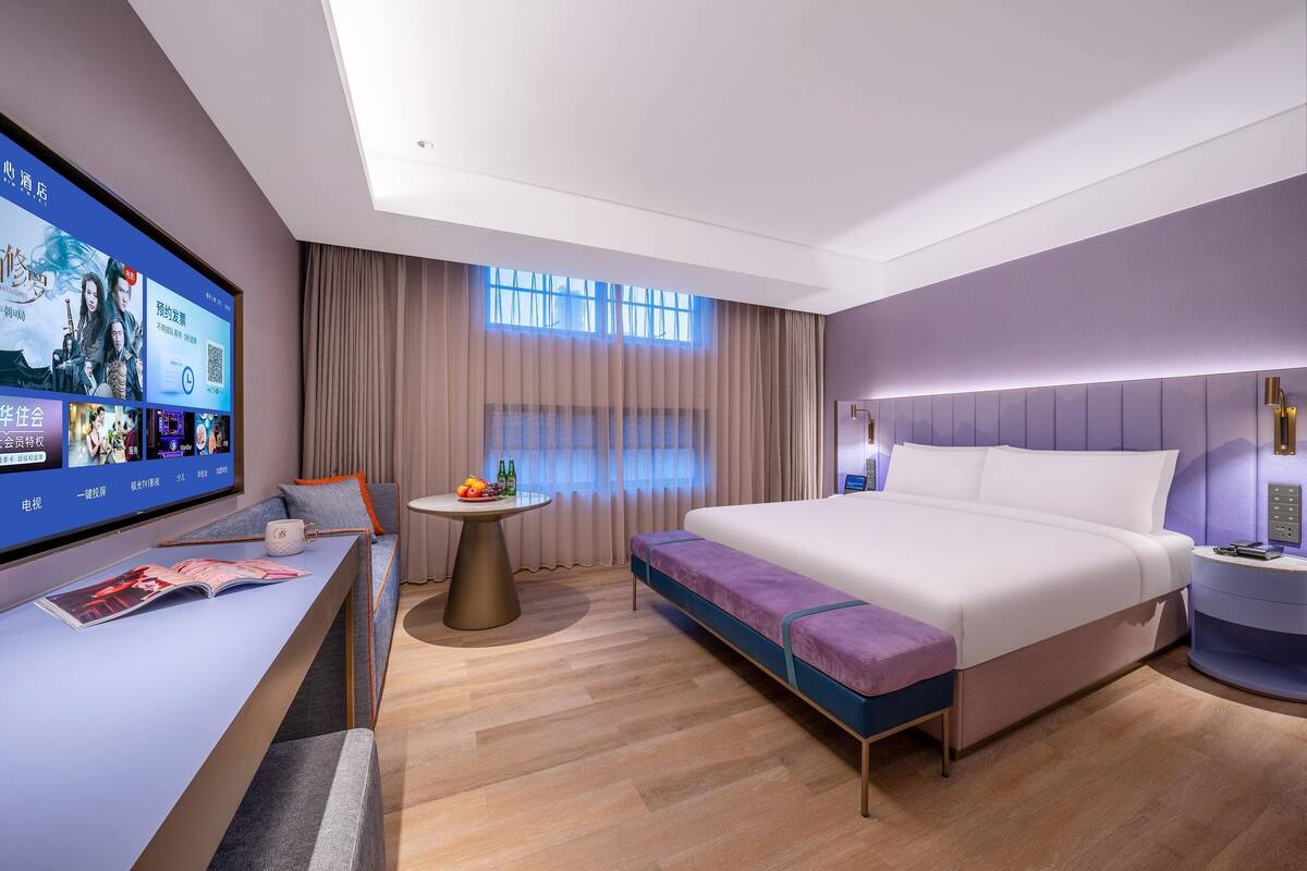 Ein modernes Hotelzimmer mit einem großen Flachbildfernseher, einer Sitzecke, einem kleinen runden Tisch mit Früchten, einem Bett mit violetten Akzenten und einem Nachttisch mit Lampe.