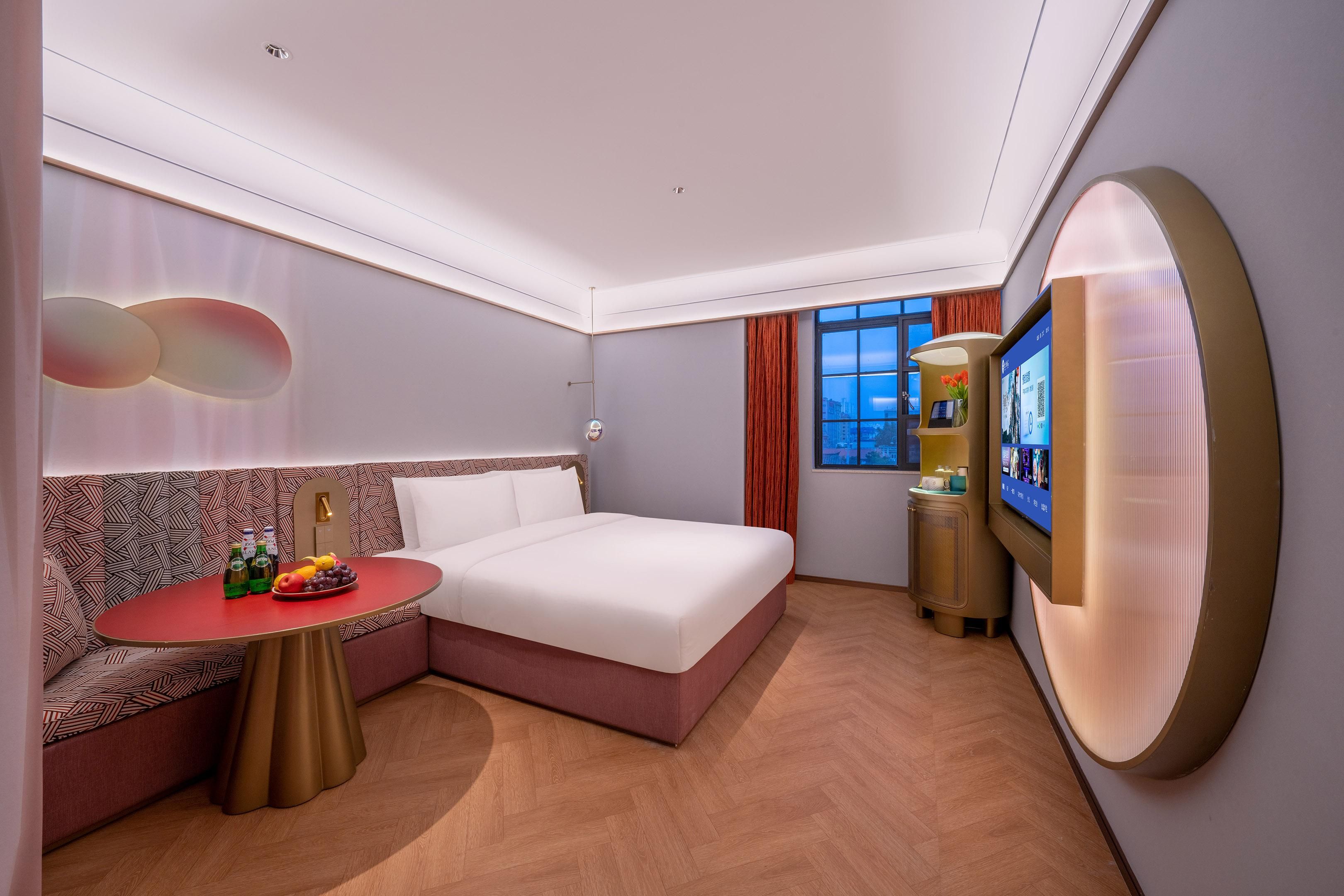 Une chambre d’hôtel moderne équipée d’un lit, d’un coin salon, d’une télévision et d’une petite table avec des boissons et des fruits.